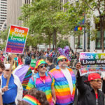 Image from a Pride protest, containing a crowd of people dressed in rainbow colours carrying rainbow placards